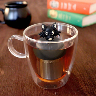 A clear double-walled glass mug filled with tea on a wooden table, featuring a black bat-shaped tea infuser and a small black bat figurine perched on top. Books are blurred in the background.