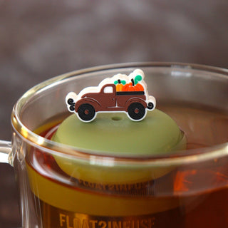 A small decorative pin shaped like a brown pickup truck carrying orange pumpkins is placed on top of a green tea infuser inside a clear glass mug filled with tea.