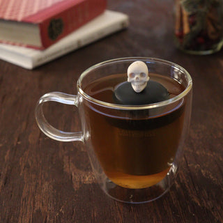 Clear glass mug with a skull-shaped tea infuser in a cup of tea on a wooden surface.