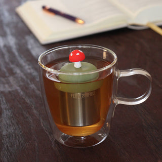 Clear mug with a mushroom-shaped tea infuser in a cup of tea on a wooden surface.